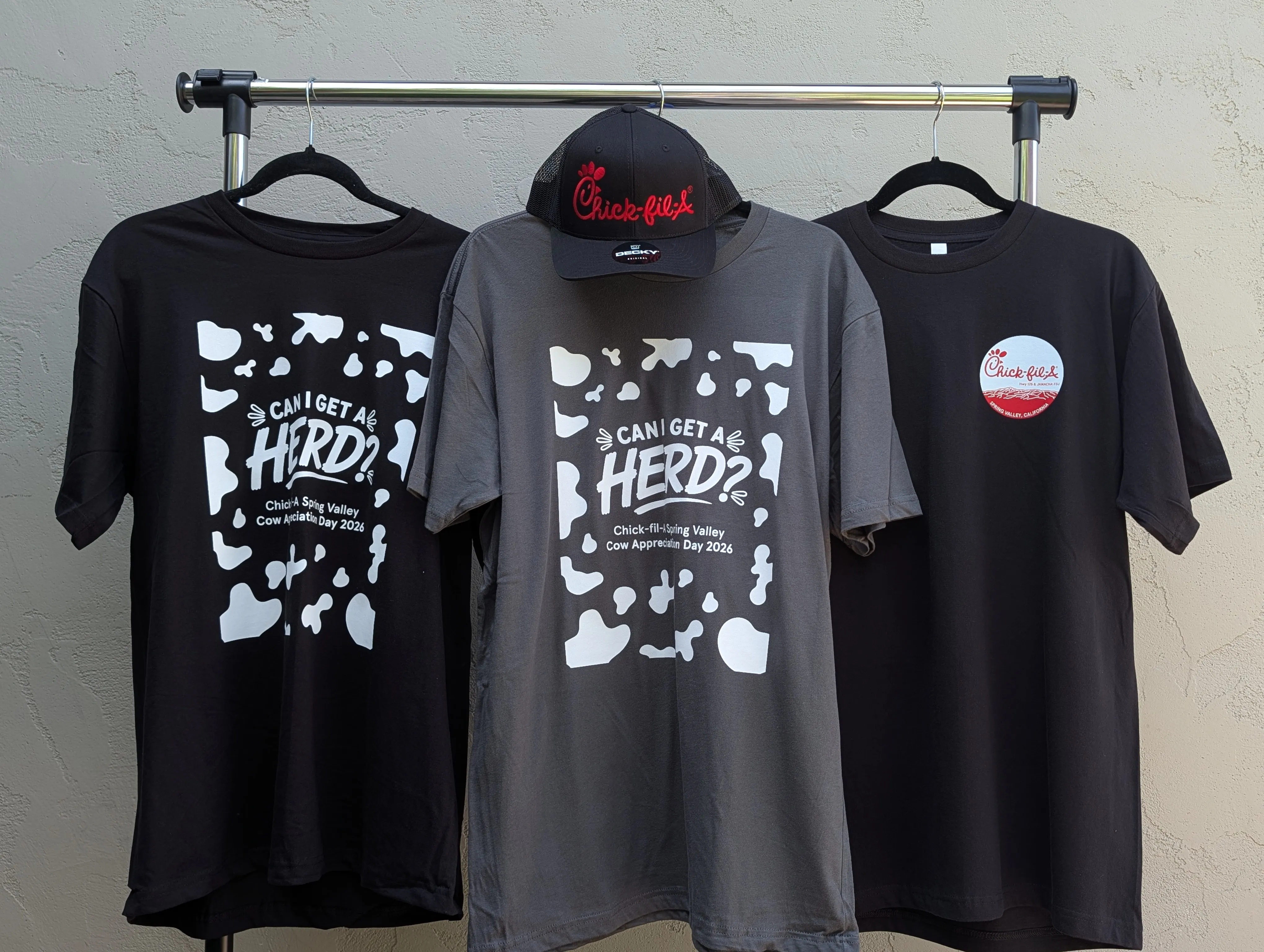 Rack of finished Cow Appreciation Day gear for Chick-fil-A Spring Valley, with cow-print 'Can I get a herd?' staff tees in black and charcoal, a black giveaway tee with the location's circle logo, and an embroidered Chick-fil-A trucker cap