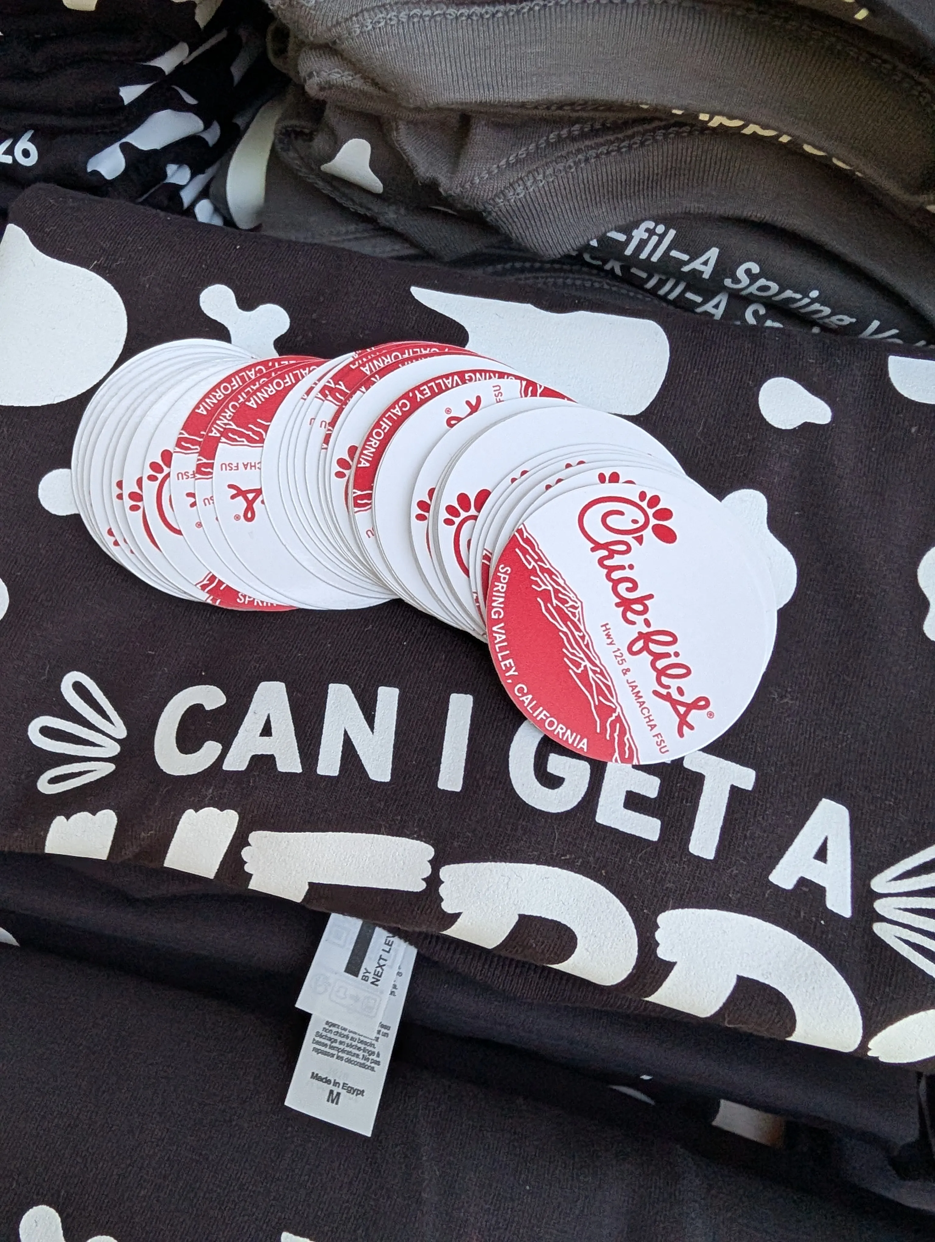 Pile of die-cut Chick-fil-A Spring Valley giveaway stickers fanned out on top of folded staff tees