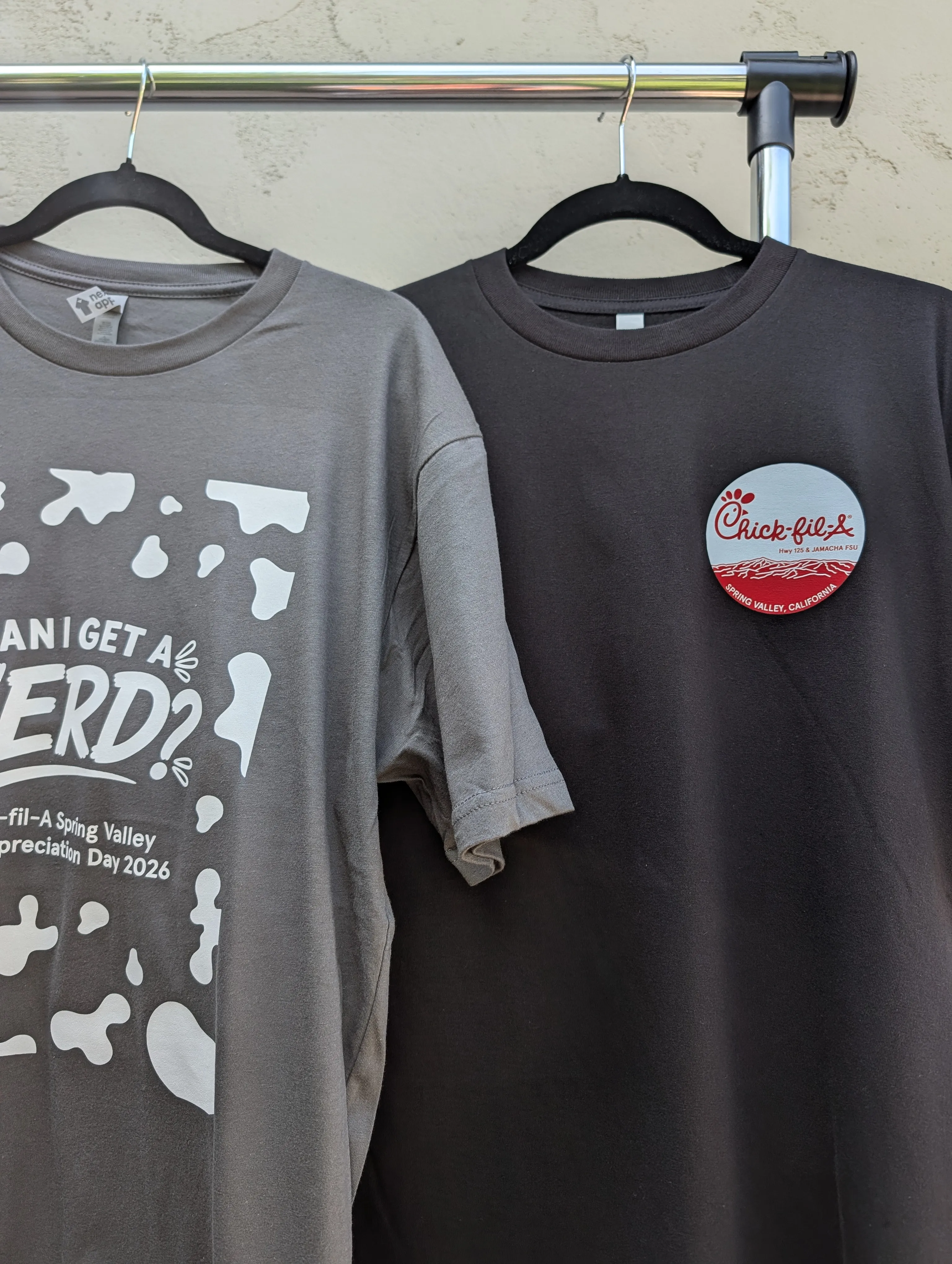 Charcoal cow-print 'Can I get a herd?' staff tee hanging beside a black giveaway tee with the Chick-fil-A Spring Valley circle logo